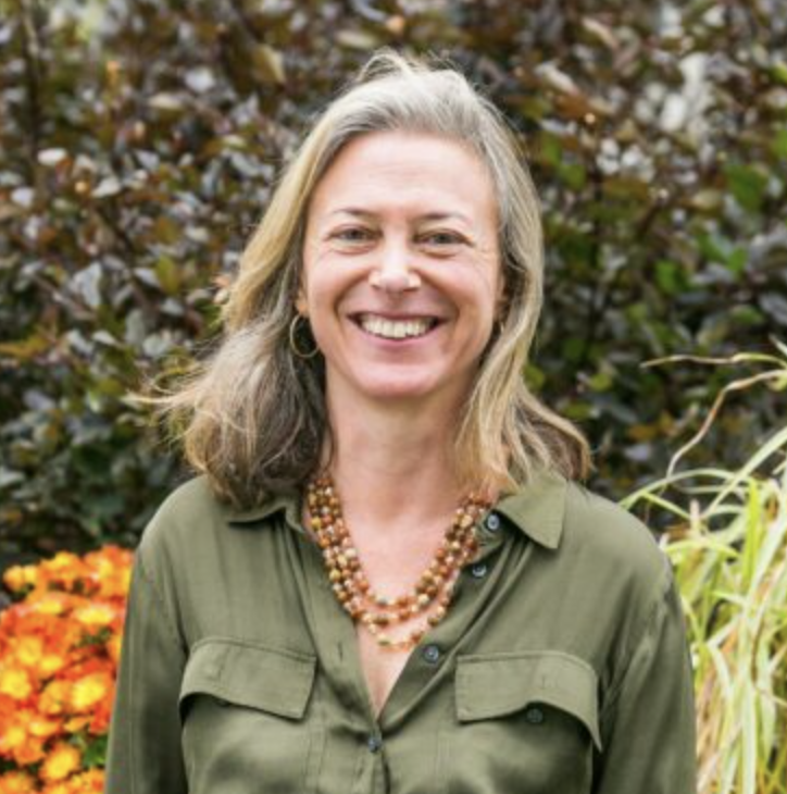 A smiling woman with shoulder-length gray hair, wearing a green button-up shirt and a multi-strand necklace, standing outdoors with autumn foliage and flowers in the background.