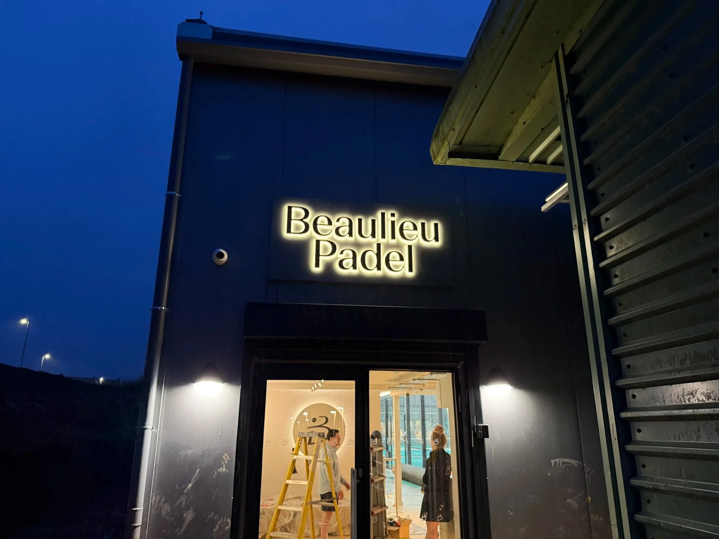 Building with illuminated sign that reads 'Beaulieu Padel' at dusk, with an open door revealing people working inside and construction materials.