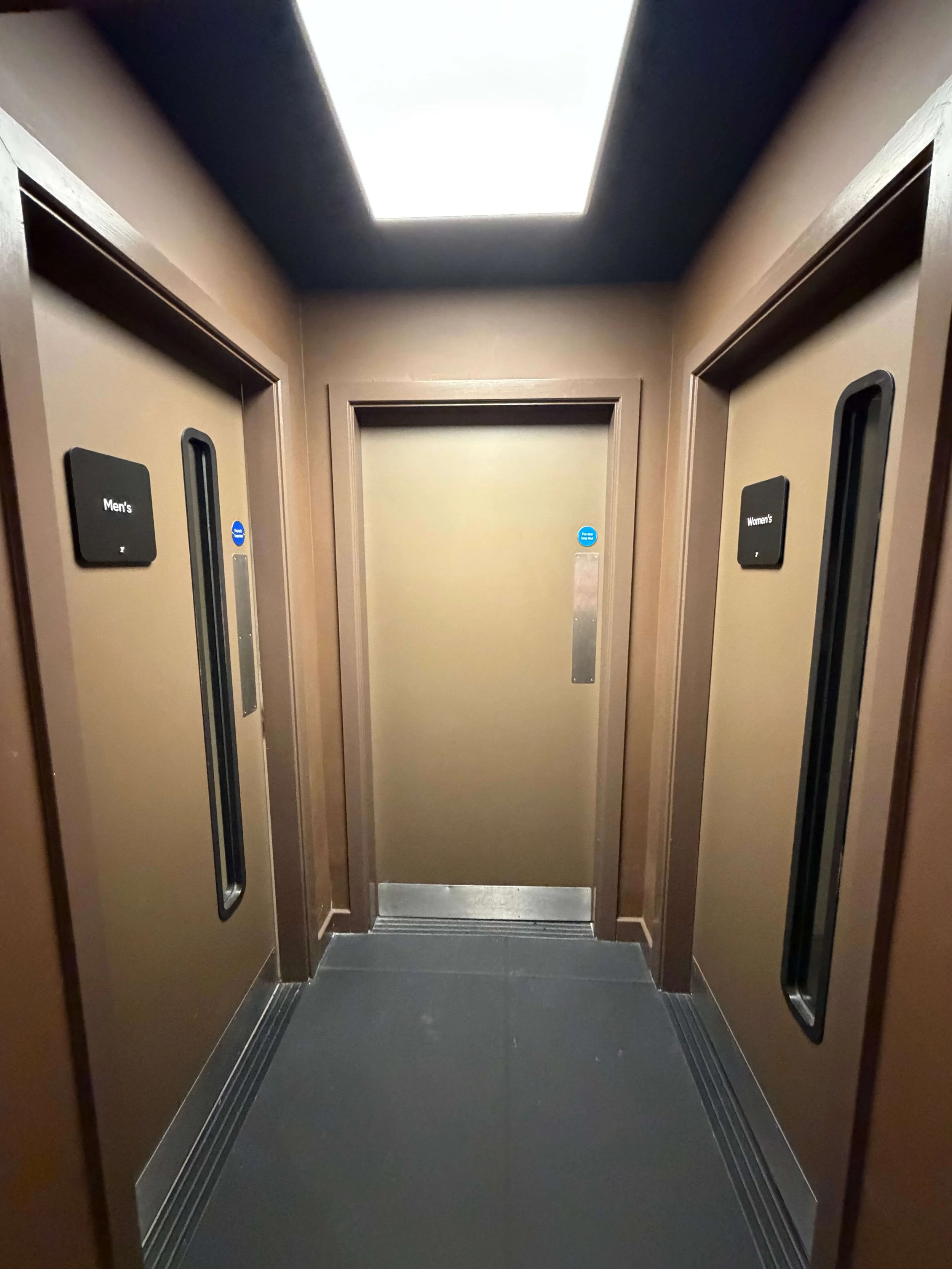 Elevator interior with closed doors labeled Men's and Women's restrooms on either side, ceiling light, and gray flooring.