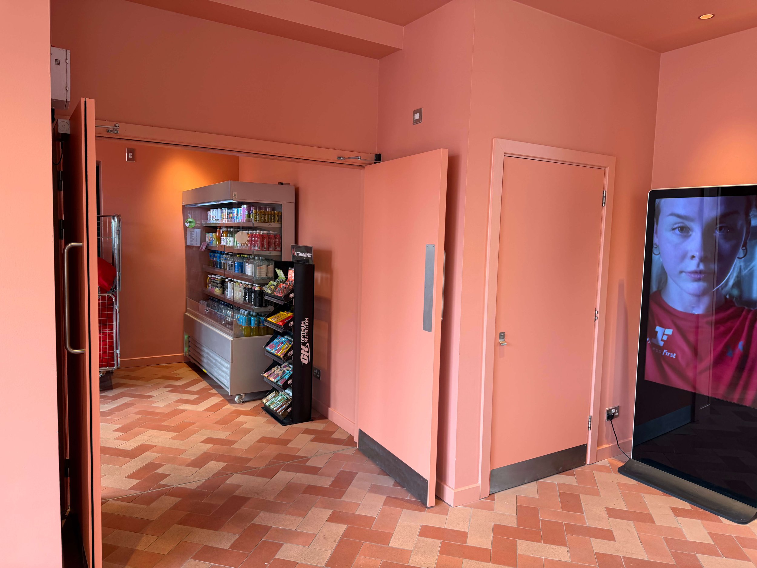 Interior view of a retail space with pink walls and brick-patterned flooring. A vending fridge and a magazine stand are visible, with part of a large digital screen showing a young girl's face on the right side of the image.