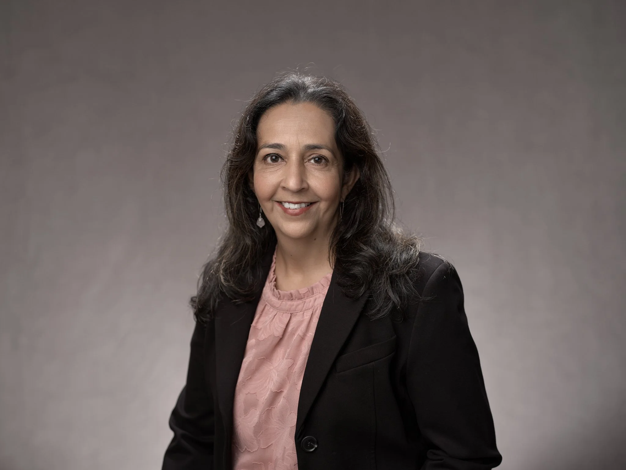 Portrait of a woman with long dark hair, wearing a black blazer and pink blouse, smiling against a plain gray background.
