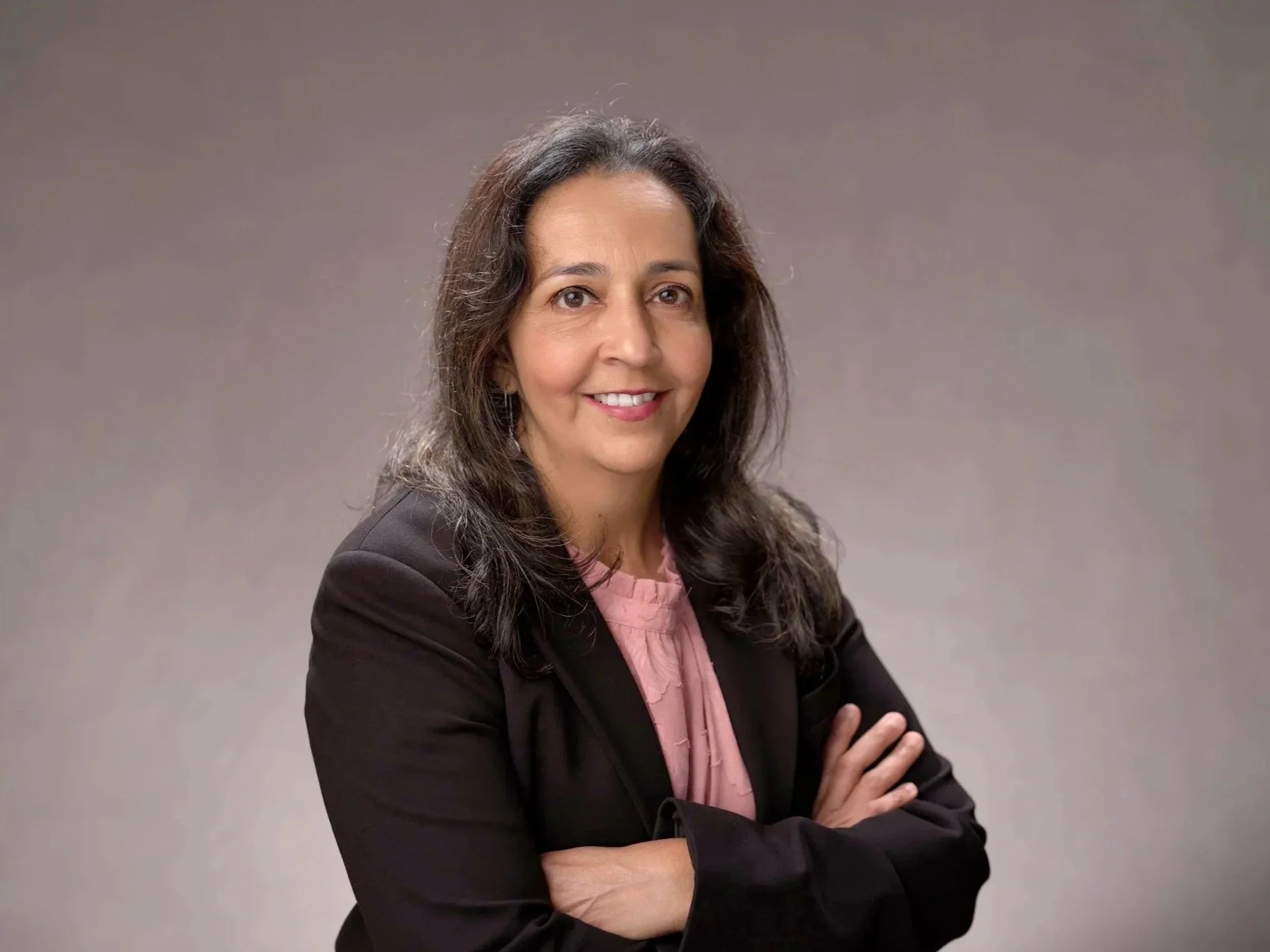 A woman with dark, wavy hair wearing a black blazer over a pink blouse, smiling with arms crossed, against a plain gray background.