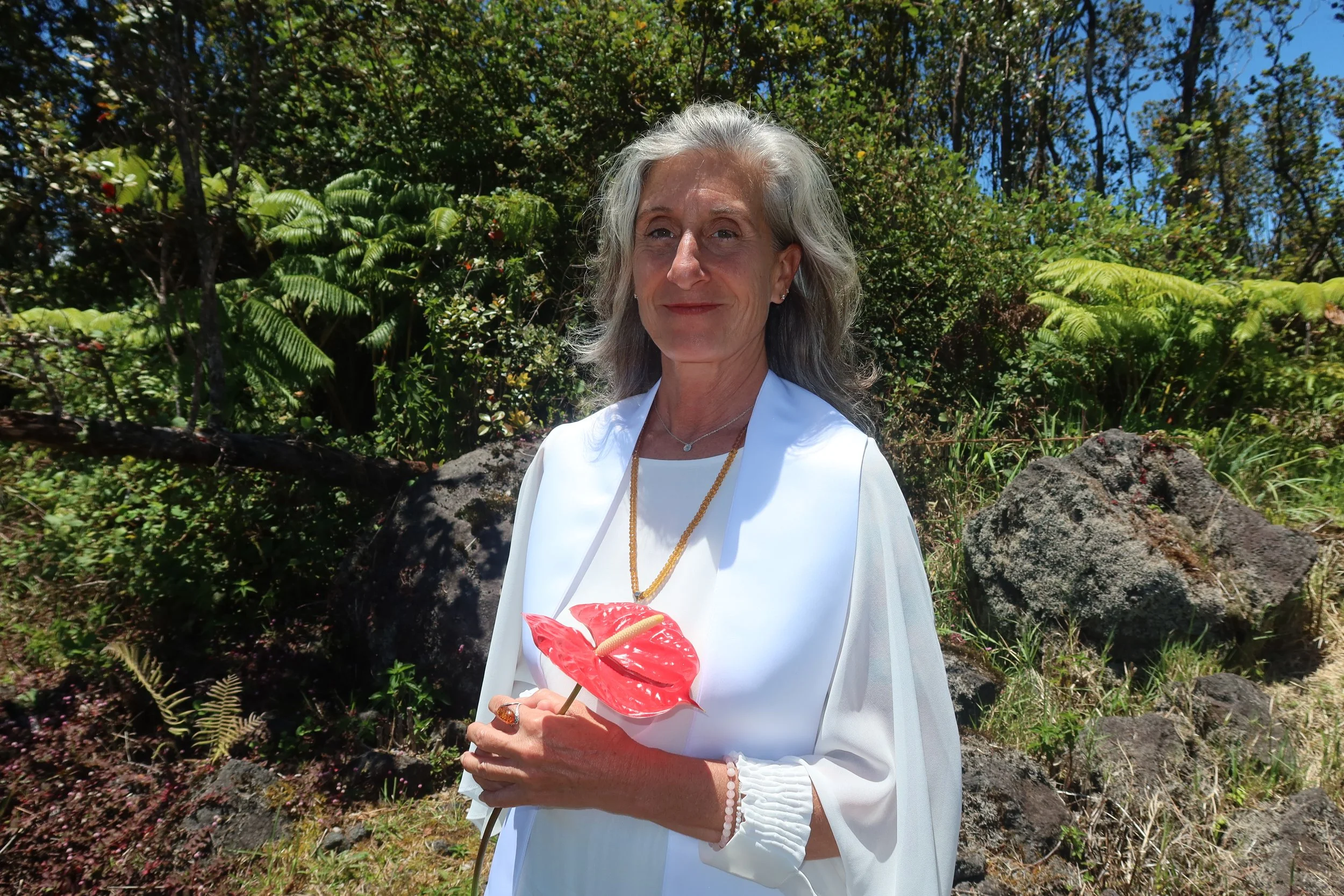 A smiling elderly woman with gray hair is standing outdoors in front of green foliage and rocks. She is wearing a white dress, jewelry, and holding a red lips-shaped paper parasol.