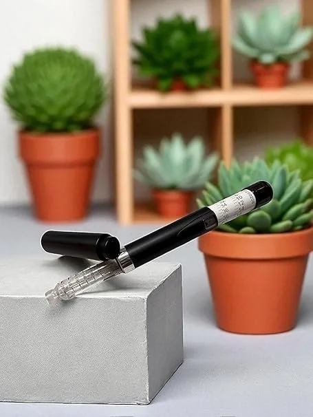 A black V2 reuseable pen and transparent barrel rests on a gray surface, with potted succulent plants and a wooden shelf in the background.