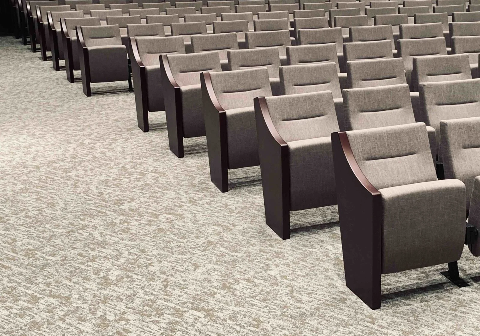Multiple rows of gray upholstered theater or conference chairs with wooden armrests on a beige and gray patterned carpeted floor.