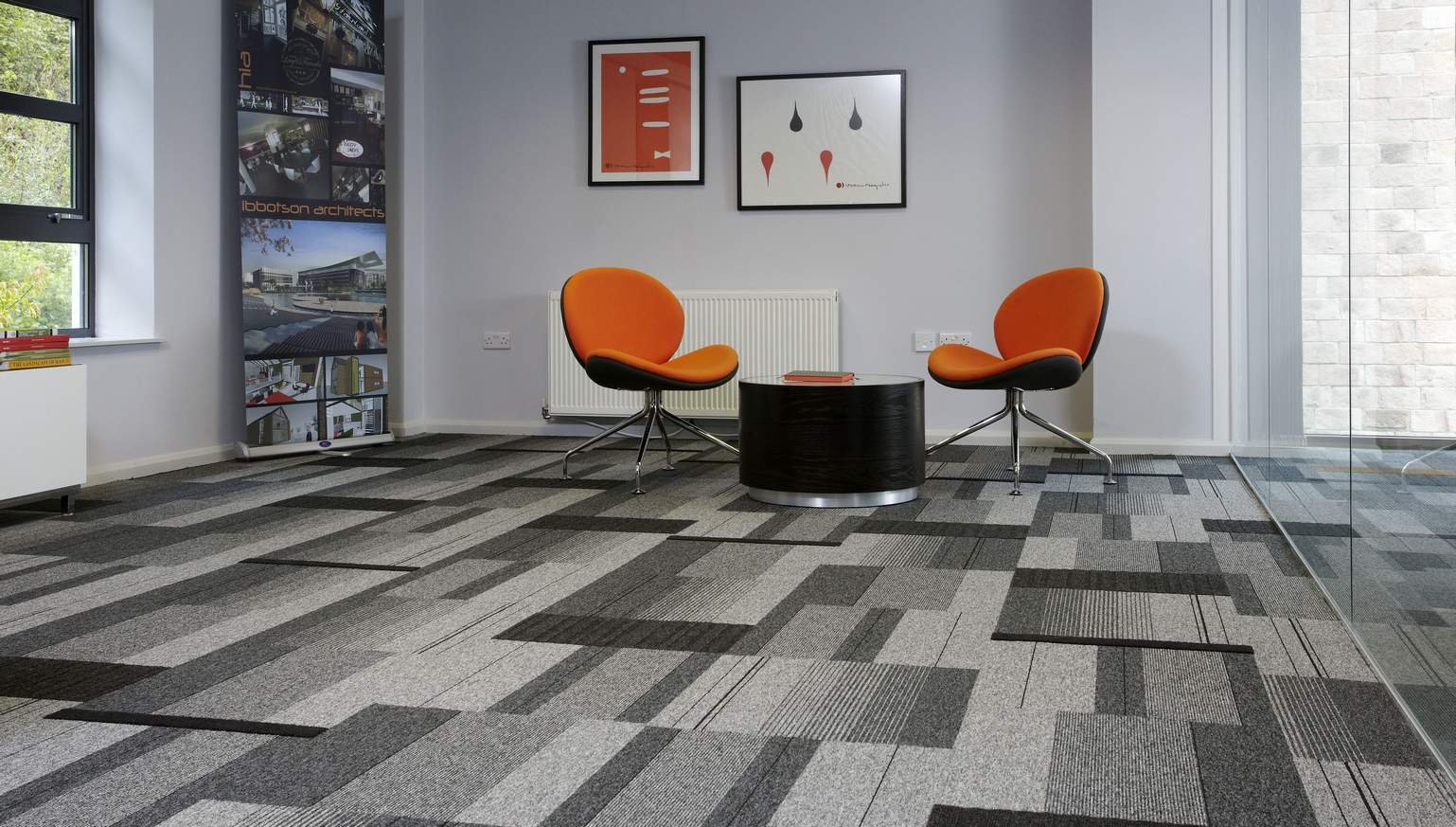 Modern office waiting area with two orange chairs, a black round coffee table, framed abstract artwork on the white wall, a large window with black frame, and patterned carpet flooring.