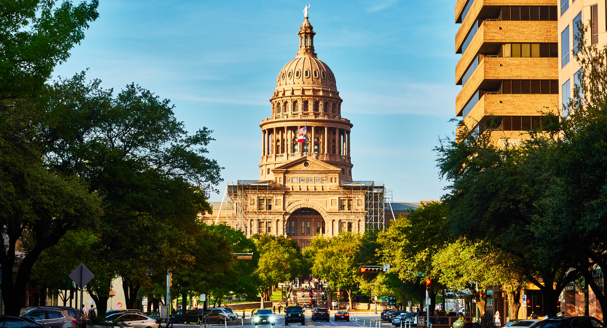 The Texas State Capitol building in Austin, Texas, with a clear blue sky, surrounded by green trees, modern buildings, and cars on the street in the foreground.