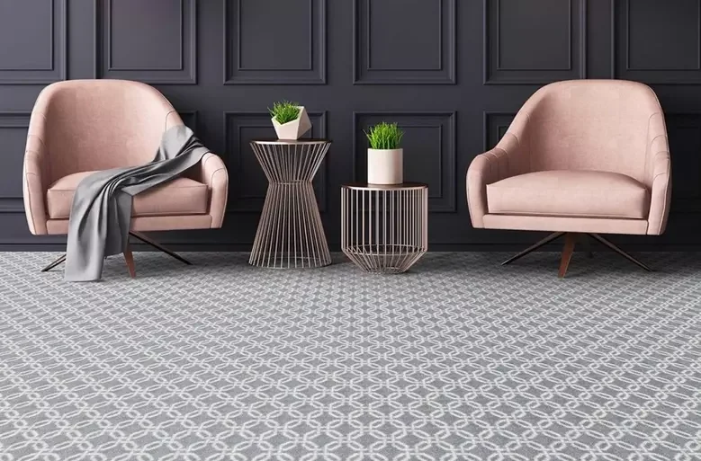 Two pink armchairs with wooden legs, one with a gray throw blanket, separated by two small round metal side tables with potted green plants, against a dark gray paneled wall on a patterned gray rug.