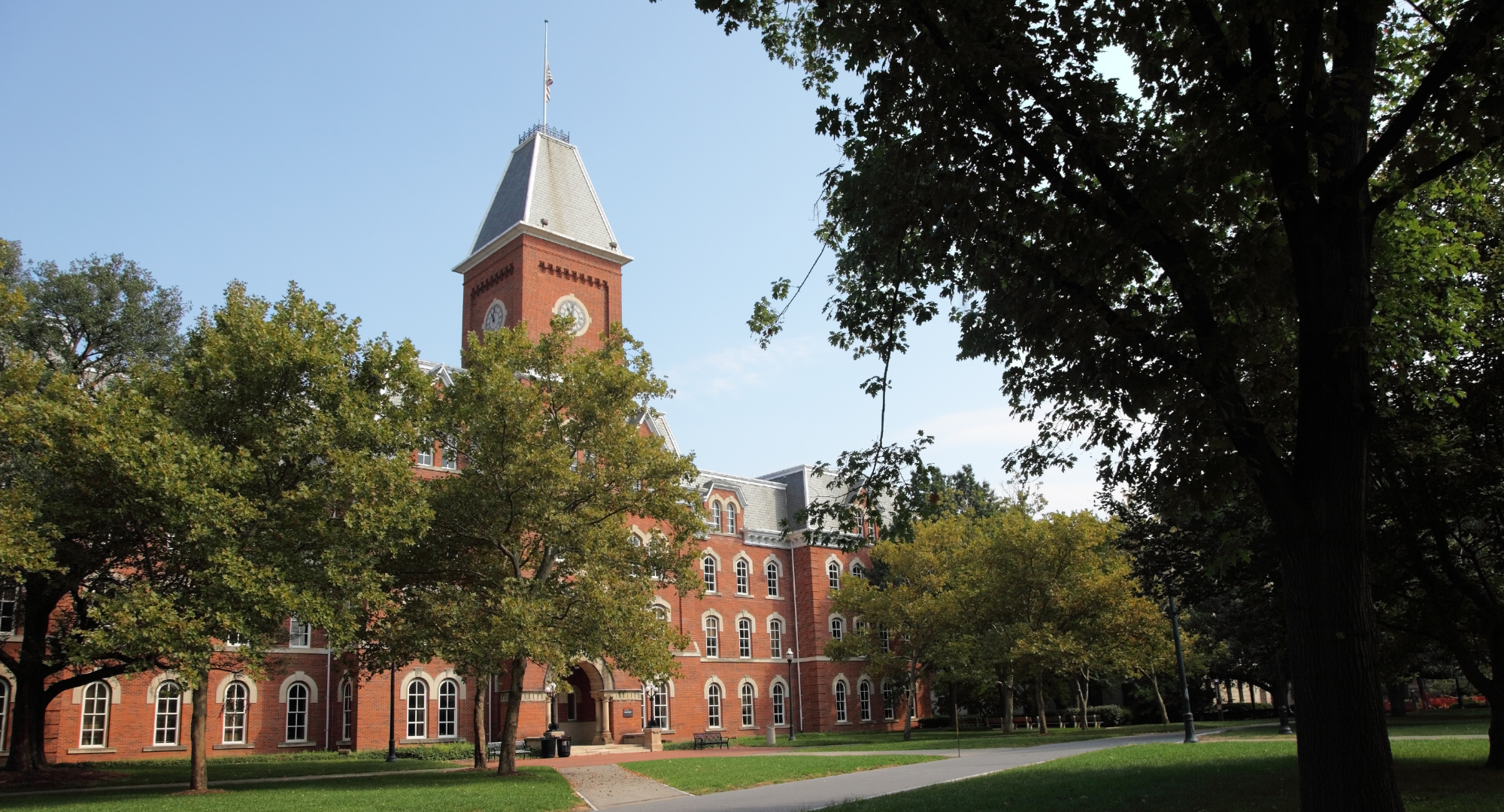 A historic red brick building with a clock tower historic building on a college campus, trees and a university lawn in foreground, clear blue sky.