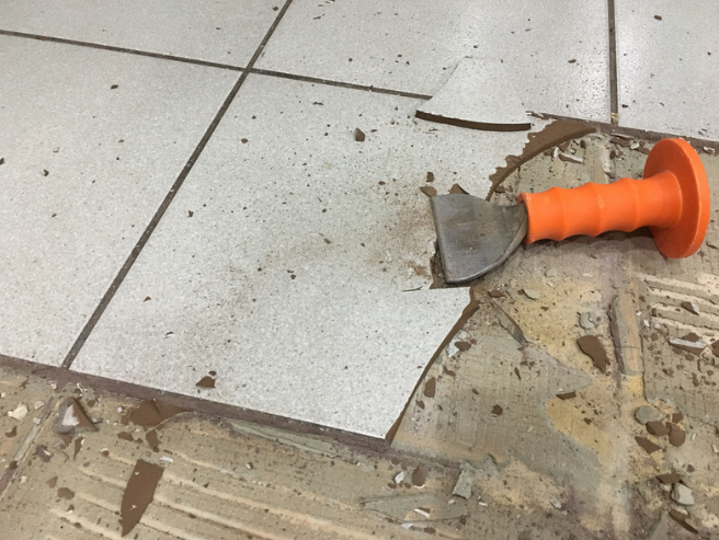 A chisel with an orange handle resting on broken beige tile with grout and debris around the broken area.