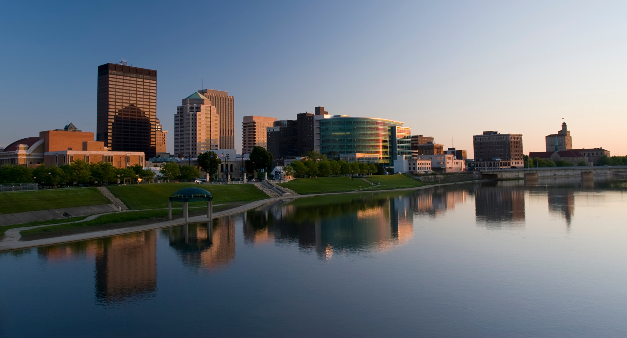 City skyline with tall buildings reflecting in a river during sunset, with green park area along the riverbank.