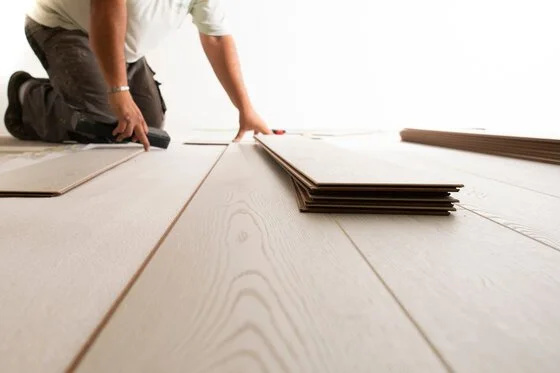 Person installing light-colored wooden flooring with stacked planks nearby.