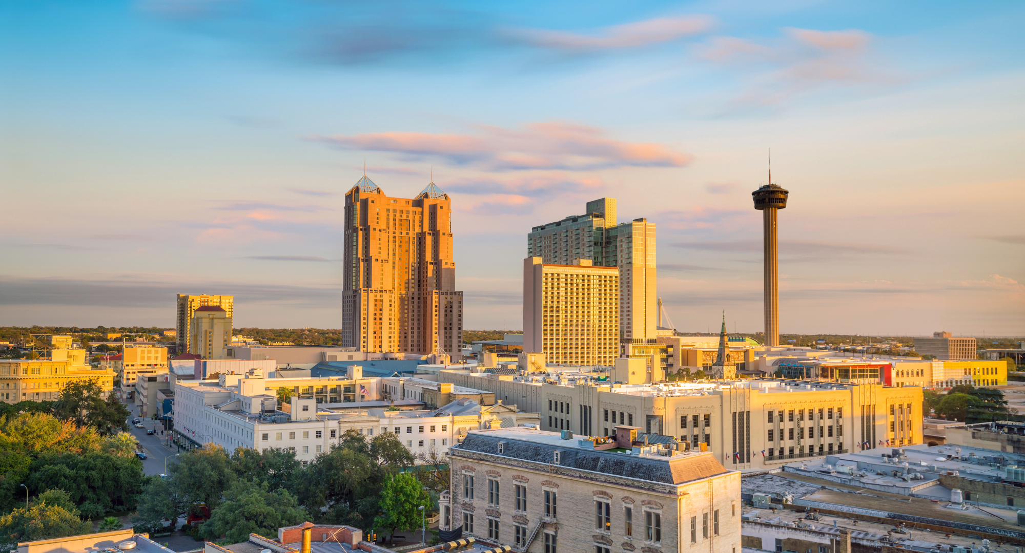 City skyline at sunset with high-rise buildings, a observation tower, and trees in the foreground.