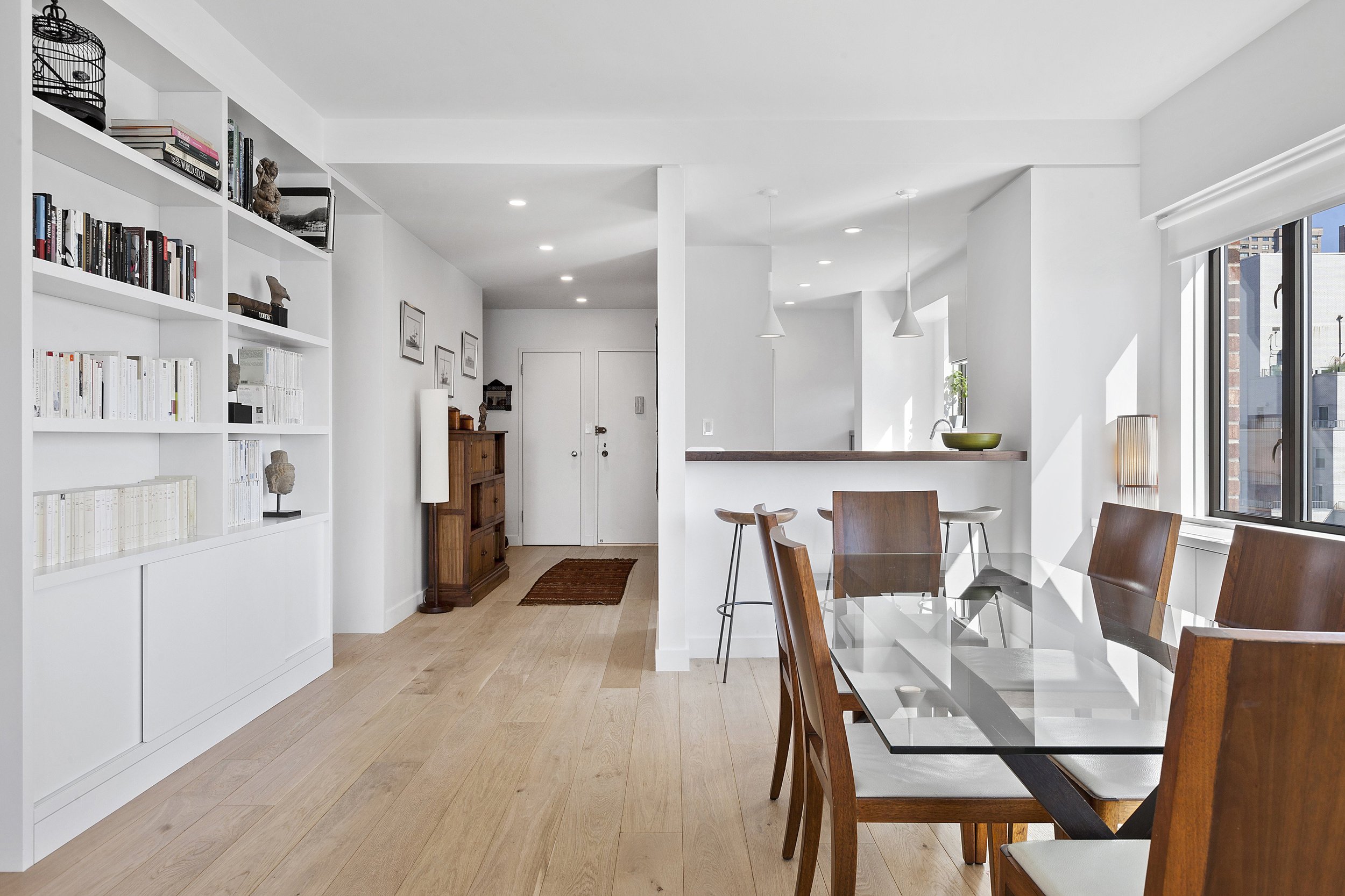 Modern dining area with a glass-top table and wooden chairs, a white wall with a large window, and a built-in bookshelf filled with books and decorations.