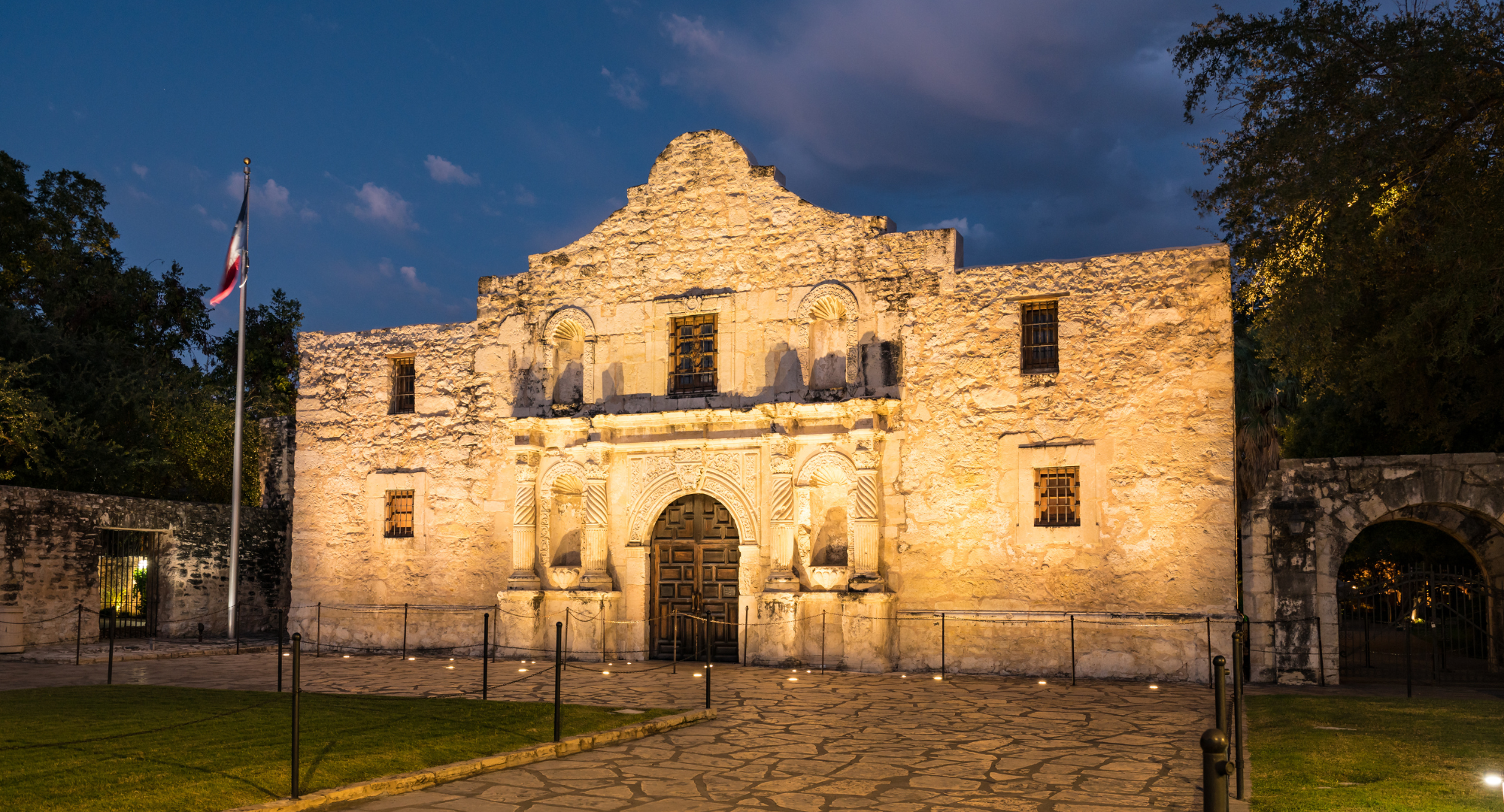 Historical stone building illuminated at night with a flagpole and a Texas flag, surrounded by trees and a paved courtyard.