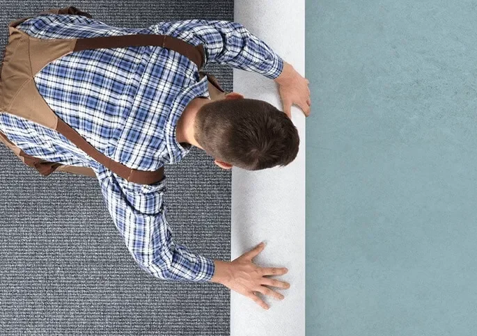 A person wearing a blue checkered shirt and khaki pants is on the floor, reaching out with their left hand, nose close to the ground, in a room with blue and gray walls.