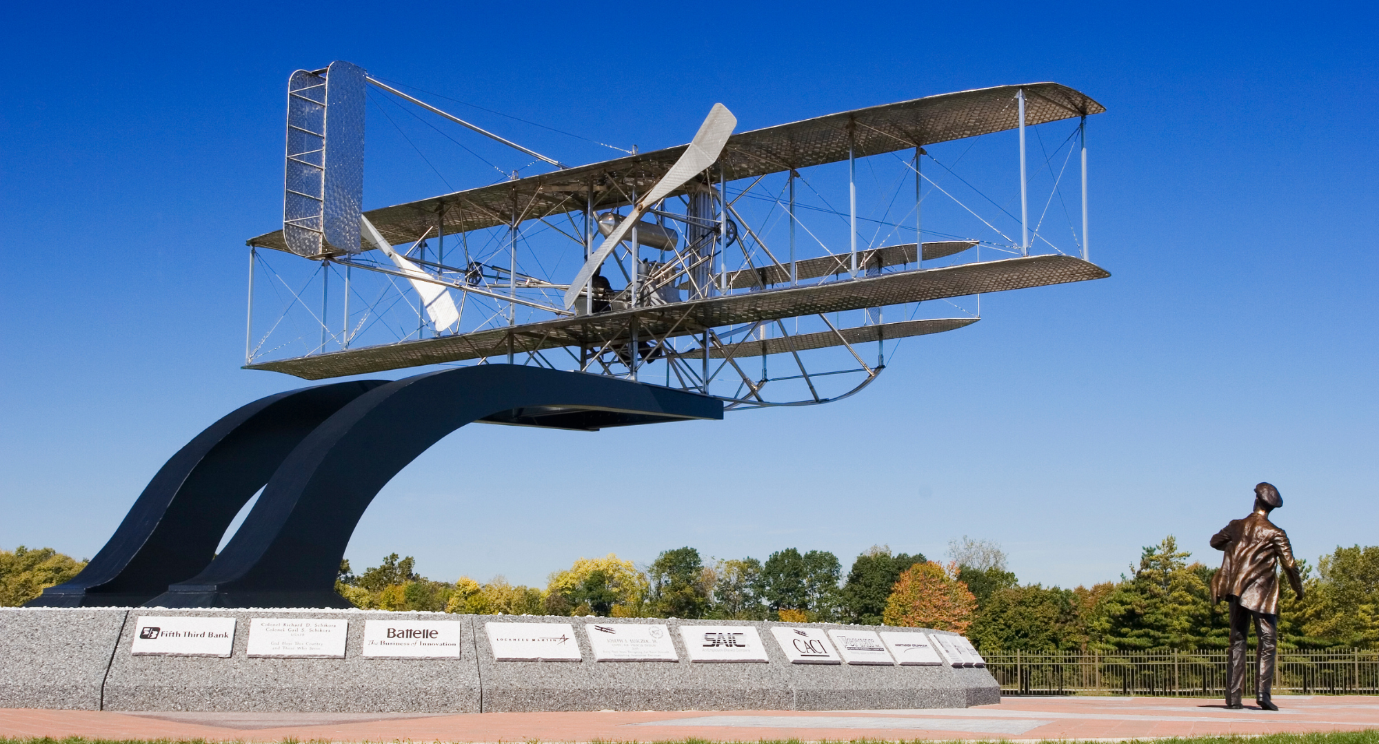 A large metallic biplane sculpture mounted on a curved black stand with an inscription plaque at its base. There is a bronze statue of a man in a coat and hat to the right, facing the sculpture. The scene is set outdoors with trees and a clear blue sky in the background.