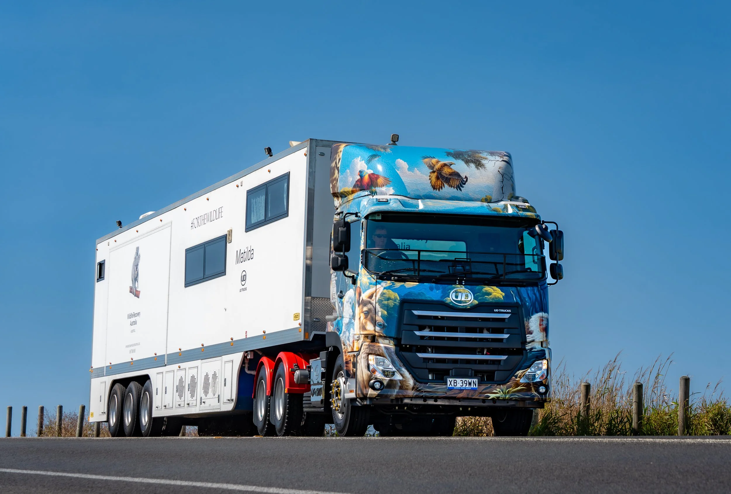 Australia's first and only mobile wildlife hospital with a striking wildlife-themed mural on the front and side, driving on a road with dry grass and wooden posts, under a clear blue sky.