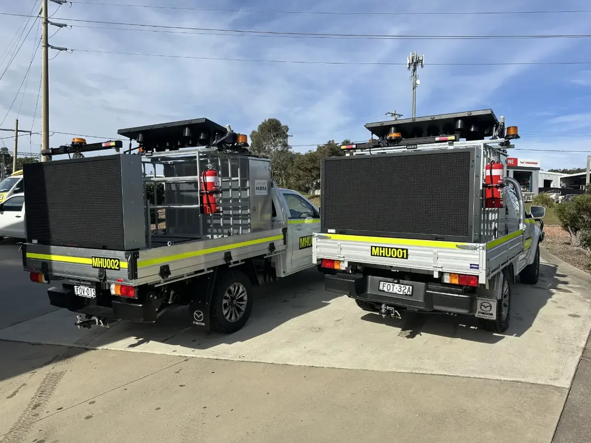 Two utility trucks with fire extinguishers and large black panels in the back, parked on a concrete surface under a blue sky.