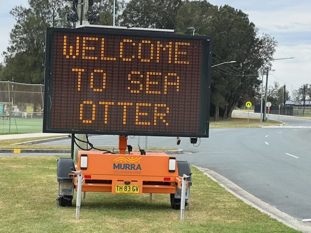 Electronic road sign on a portable stand displays the message 'WELCOME TO SEA OTTER' in yellow letters.