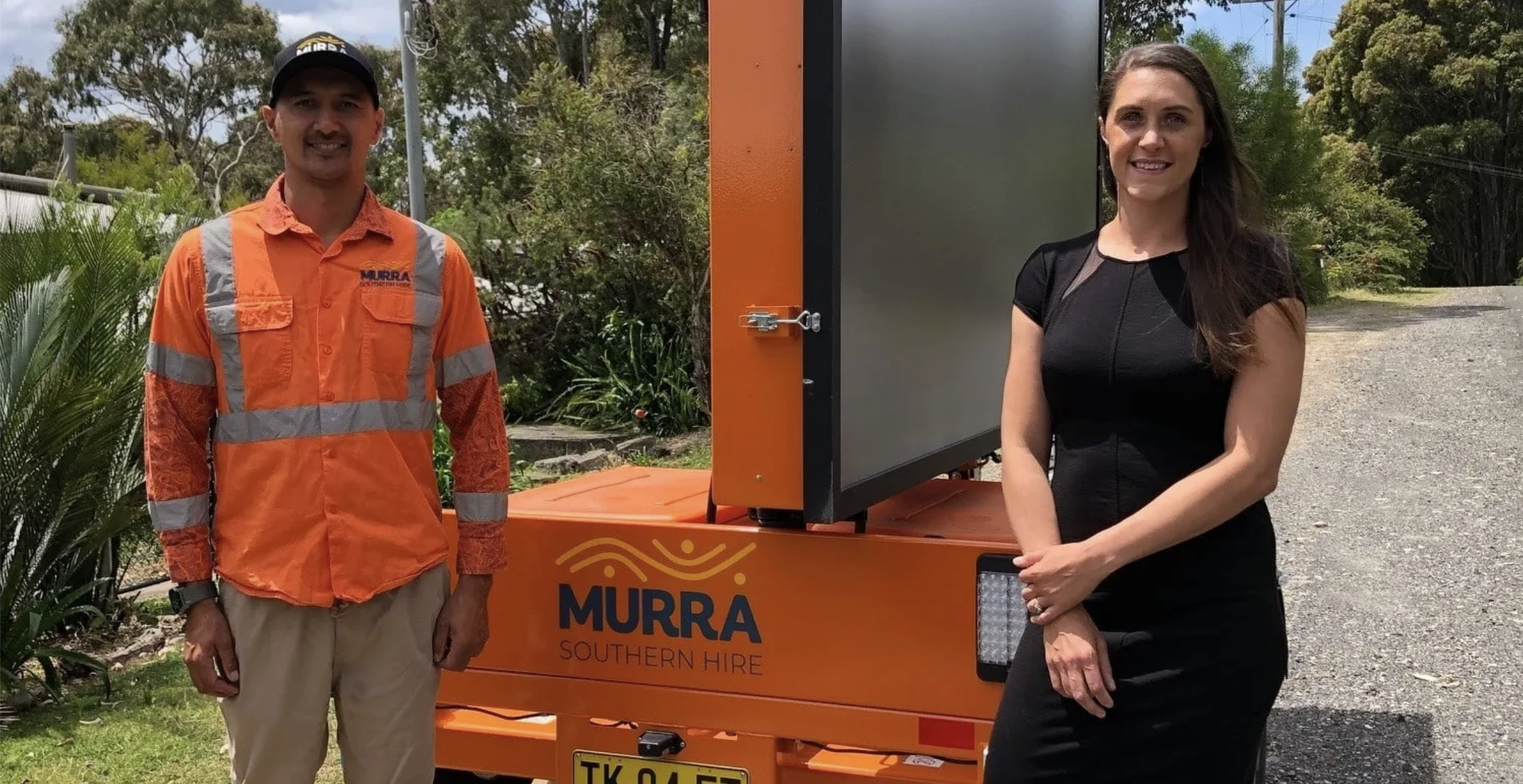 A man wearing an orange safety shirt and a cap stands next to a woman in a black dress, both smiling, outside near an orange mobile digital billboard with the sign "MURRĂ SOUTHERN HIRE". The backdrop includes greenery and a gravel path.