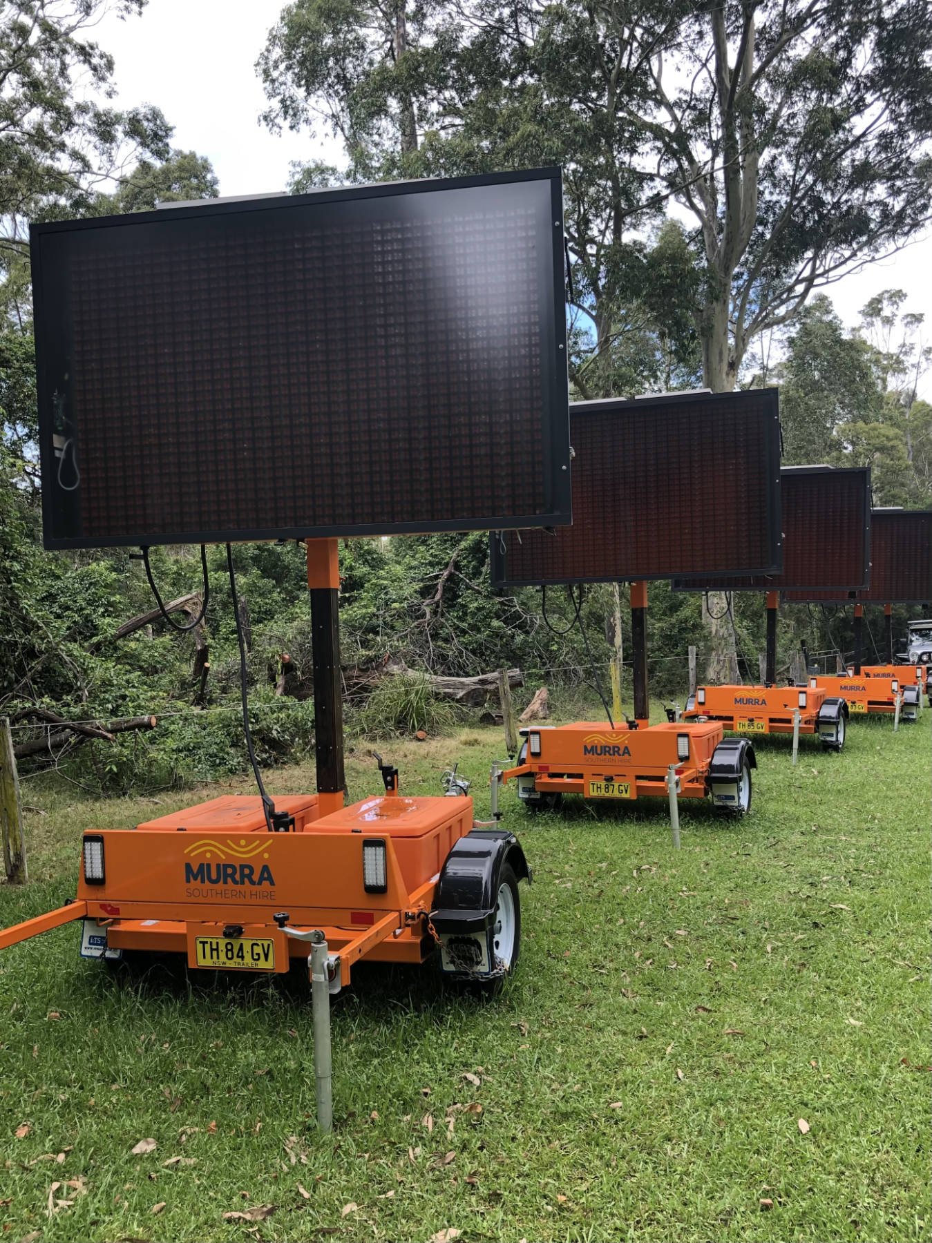 Multiple mobile electronic billboard signs mounted on orange trailers, lined up outdoors on grass with trees in the background.