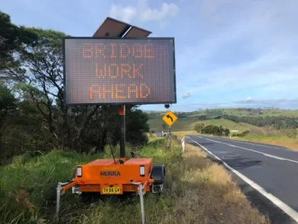 Digital roadside sign reading 'BRIDGE WORK AHEAD' near a two-lane road, with trees, grass, and hills in the background.