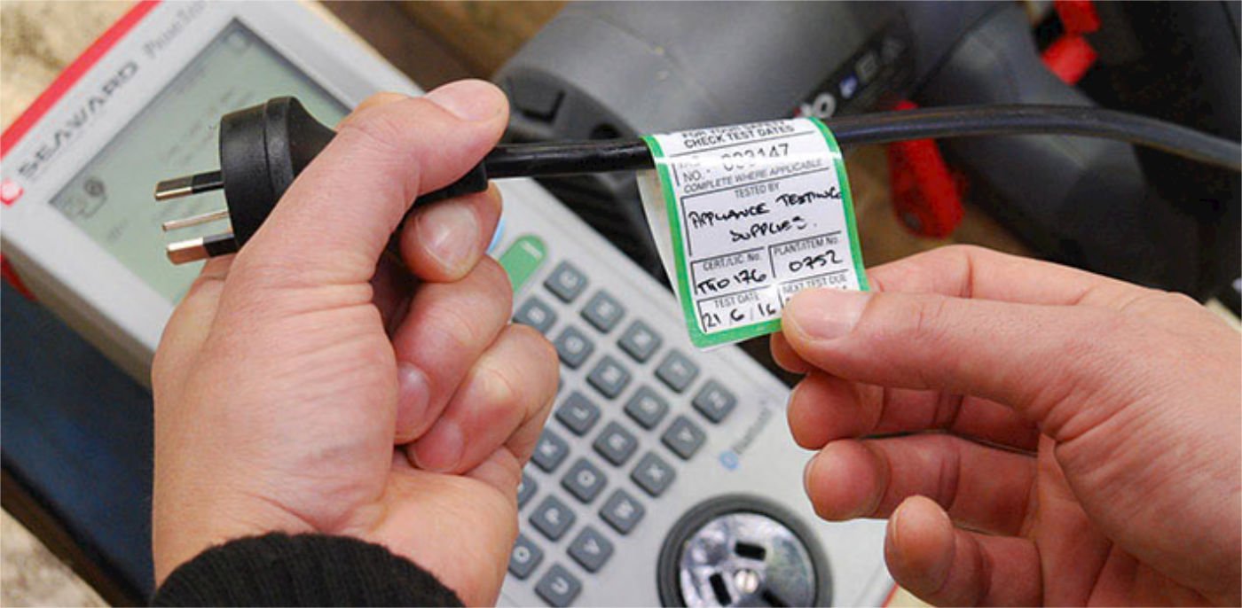 Person holding an electrical testing device with a label, in front of a multimeter, indicating electrical testing equipment.