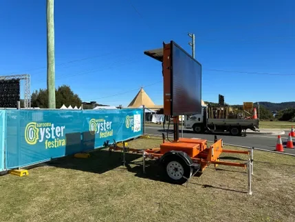 Portable electronic billboard on trailer at outdoor festival with tents and a train in the background.