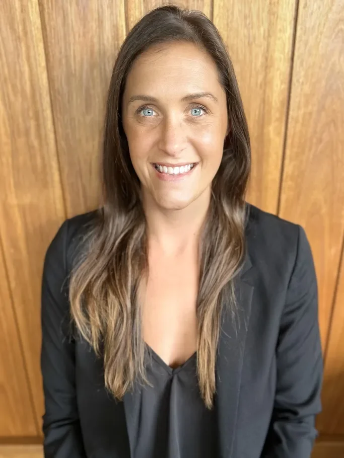 A woman with long brown hair and blue eyes, smiling and wearing a black blazer and top, standing in front of a wooden wall.