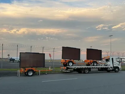 A flatbed truck transporting three large, black, rectangular billboards at an airport runway with a cloudy sky.