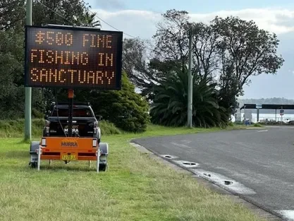 Digital sign advertising $5.00 fine for fishing in sanctuary near a road with trees and water in the background