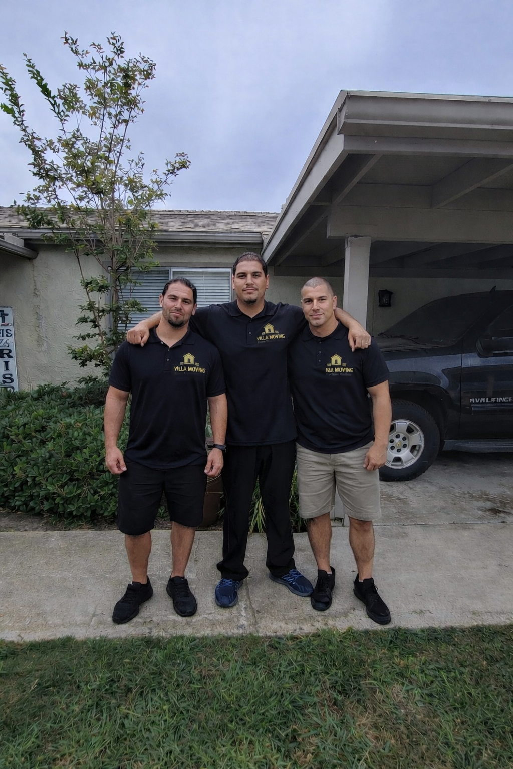 Three men standing together outside in front of a house, wearing black polo shirts with a moving company logo. The man in the middle has his arms around the shoulders of the men on either side. There is a black truck parked under a carport to the right, and a small tree and bushes to the left.