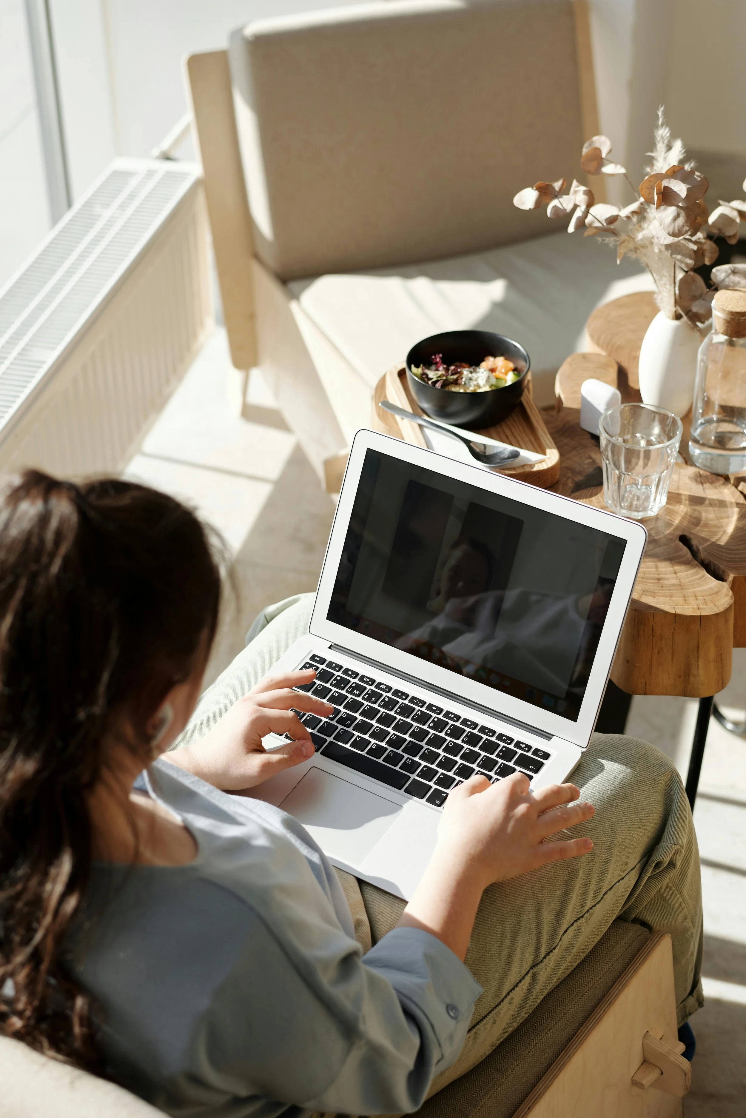 A woman sitting on a sofa using a laptop, with a wooden table nearby holding a bowl of salad, a glass of water, and a vase with dried flowers in a sunlit room.