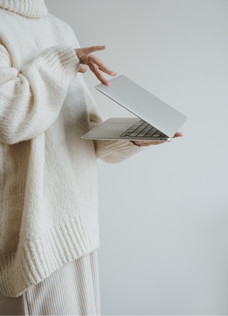 A person wearing a cream-colored knitted sweater holding a closed silver laptop with one hand and gesturing with the other hand.
