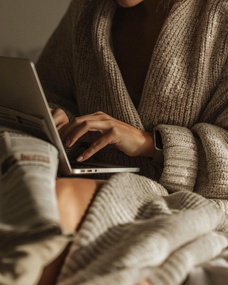 Close-up of a person wearing a chunky knit cardigan and a smartwatch, typing on a laptop with another person in a camouflage sleeve nearby.