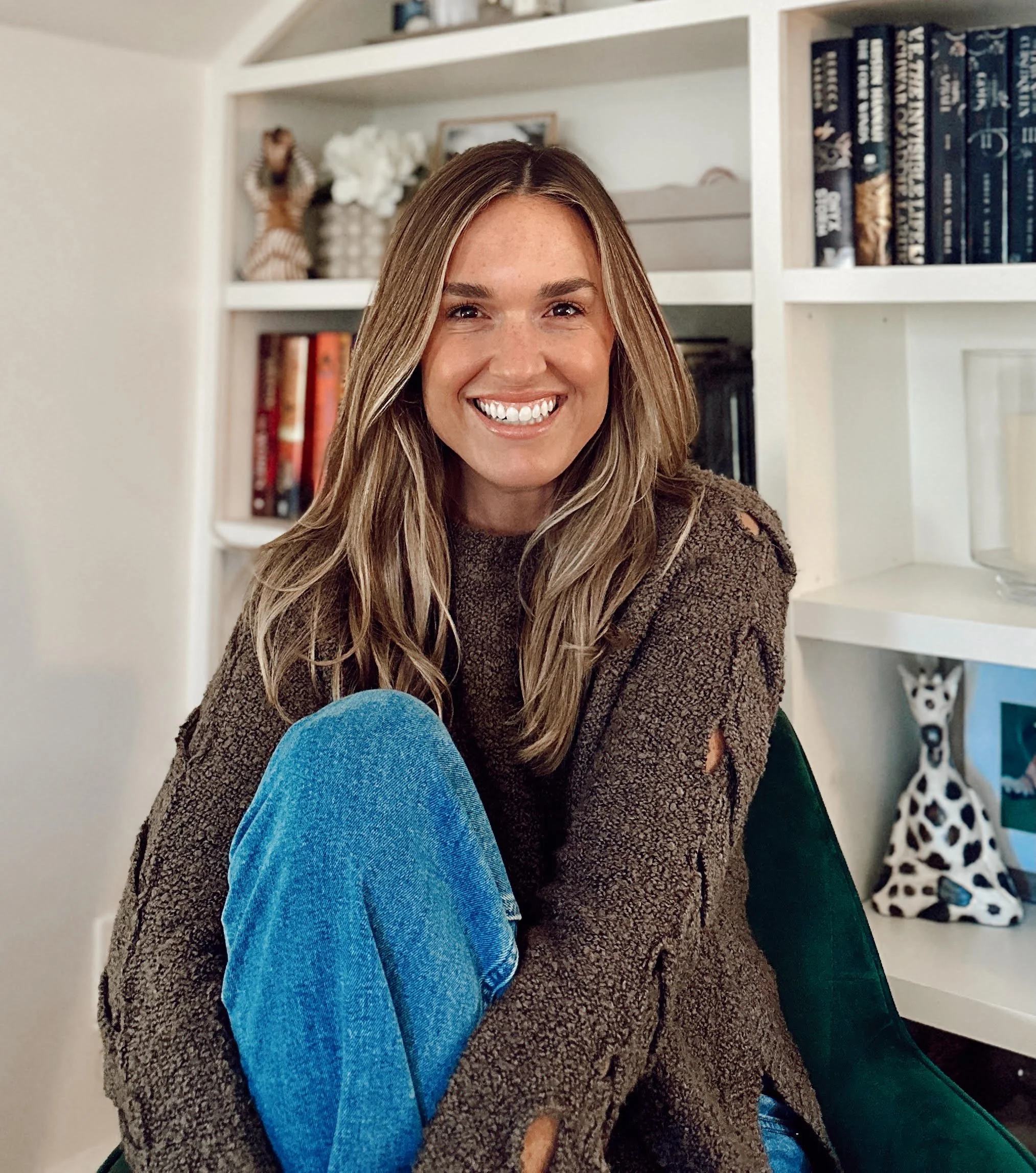A smiling woman with long, wavy blonde hair sitting on a green chair in a room with white bookshelf and decorative items.