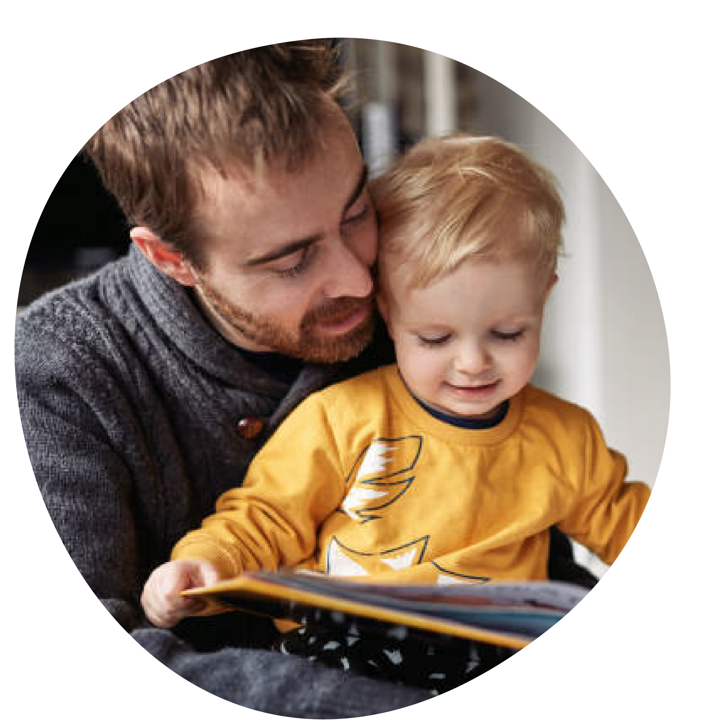 A man and a young boy, likely a father and son, are sitting together looking at a book. The man leans in closely with a gentle expression, while the boy, wearing a yellow shirt, is focused on the book.