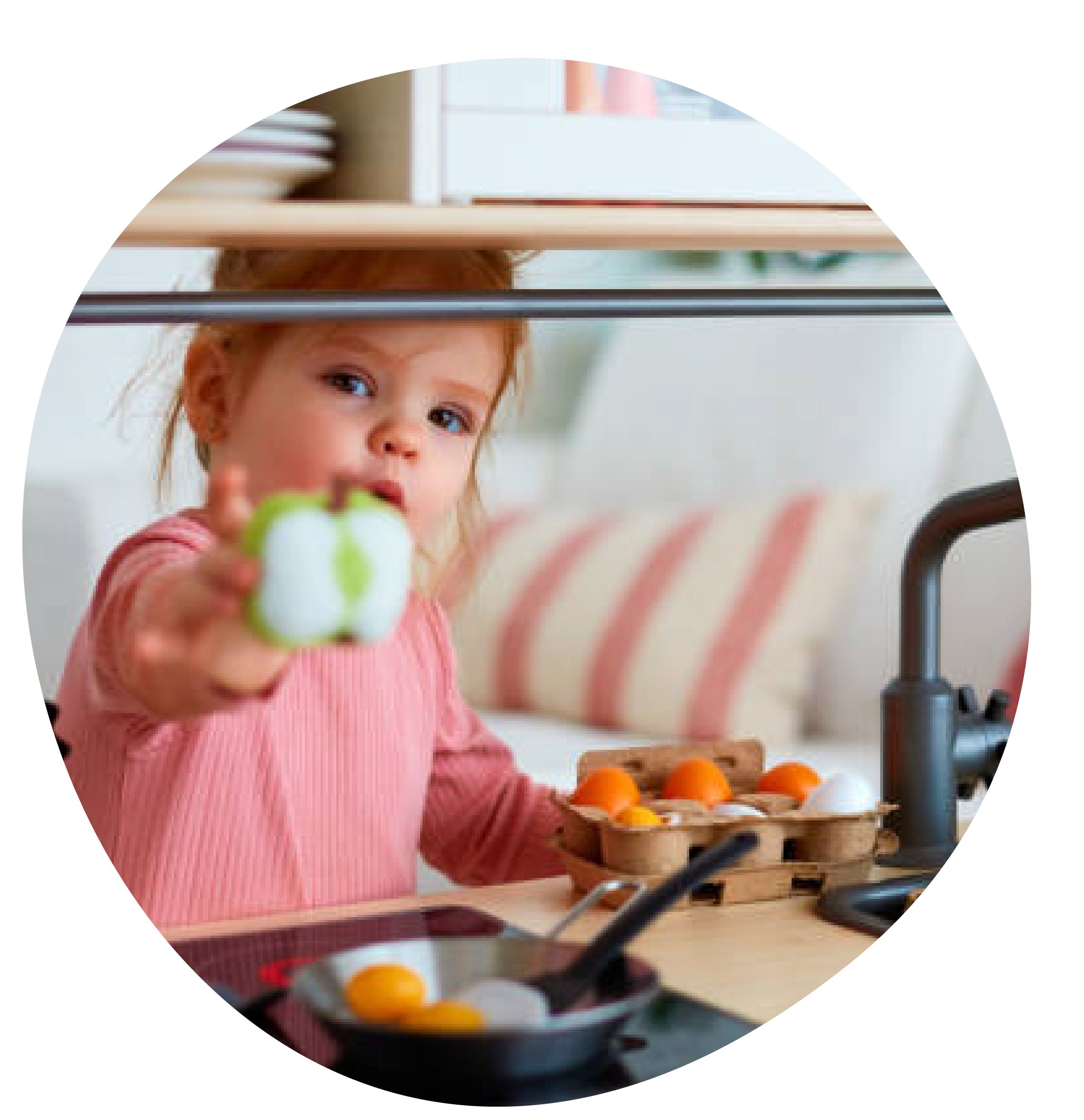 A young girl in a pink sweater holding a green and white toy apple towards the camera in a kitchen, with a carton of eggs and a frying pan on the counter.