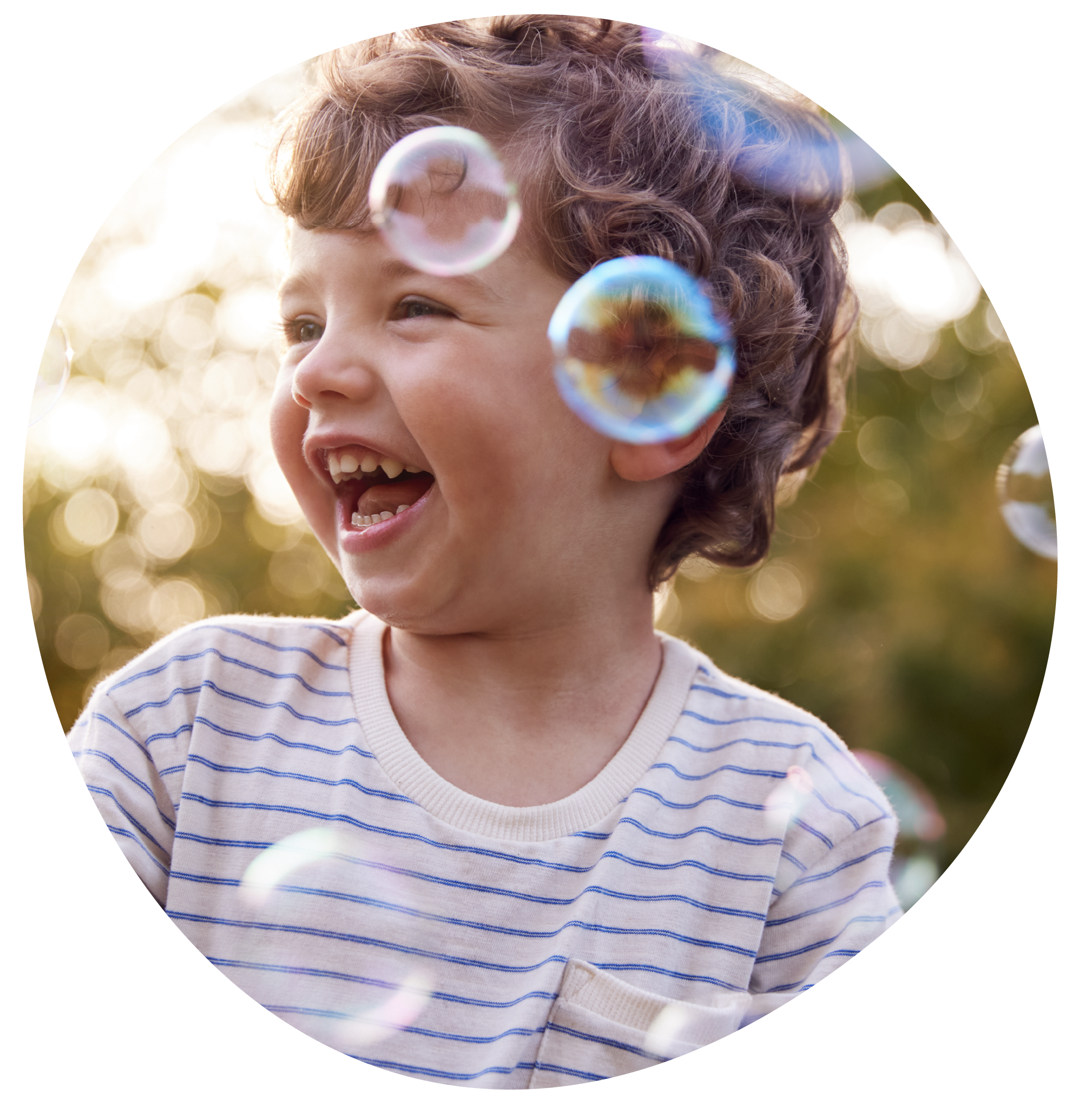 A young boy with curly brown hair smiling and playing outdoors among soap bubbles.