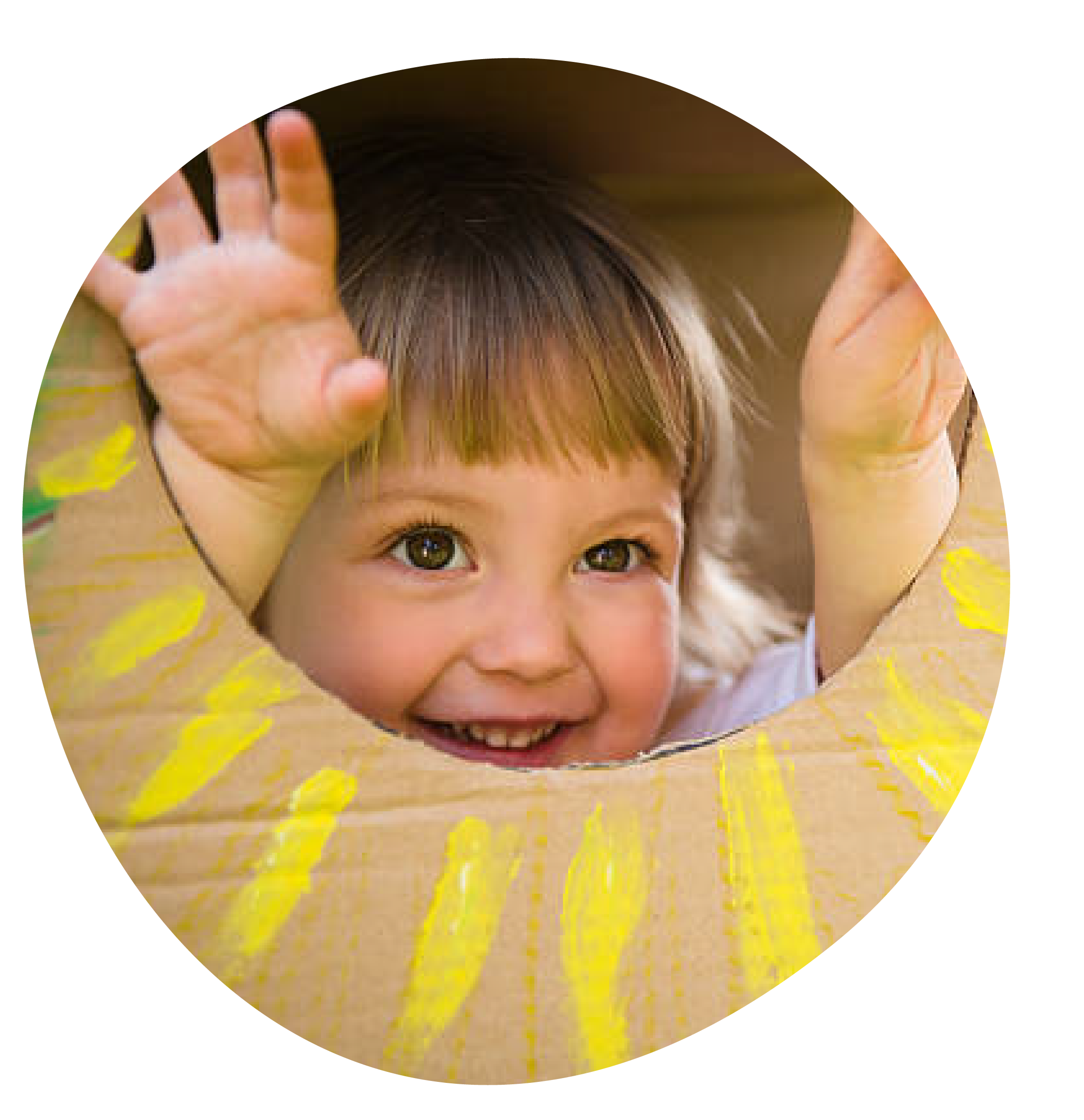 A young girl with brown hair and big brown eyes looking through a circular hole in a cardboard box, smiling and reaching her hands into the hole.