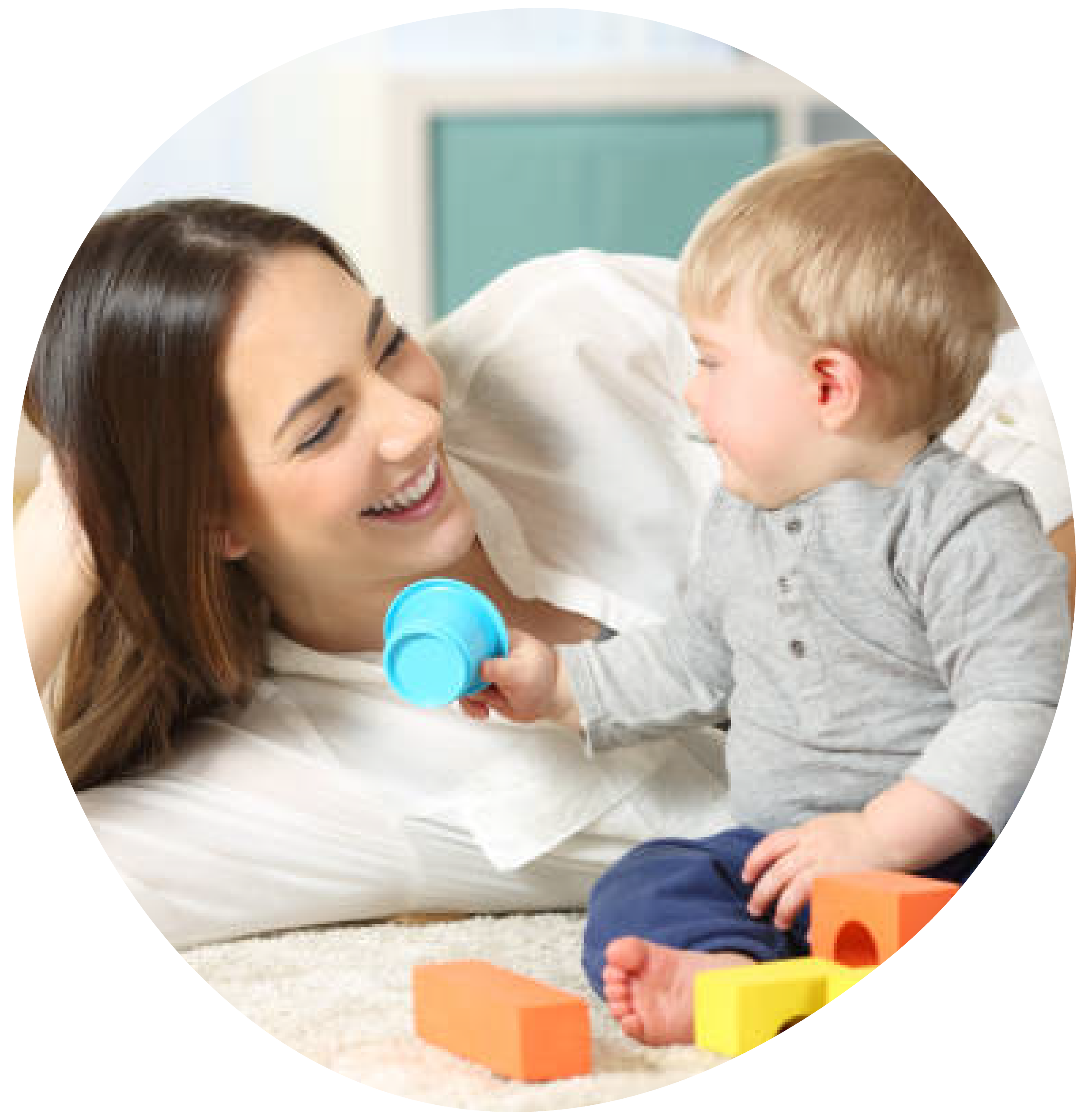 A woman and a young boy playing together on the floor of a room, smiling and enjoying themselves. The boy is holding a blue object, and there are colorful foam blocks around them.