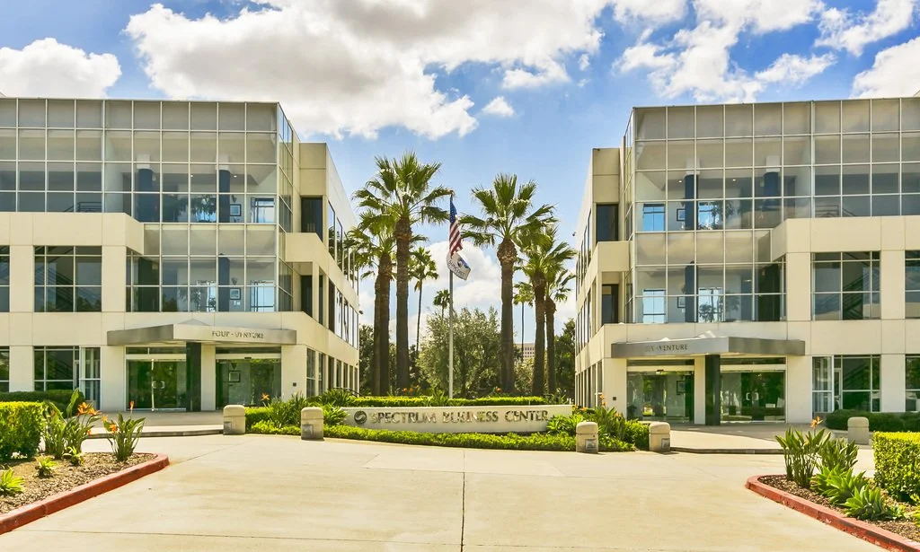 Modern office buildings with glass facades, a pathway leading to the entrance, and palm trees in the background in a sunny setting.