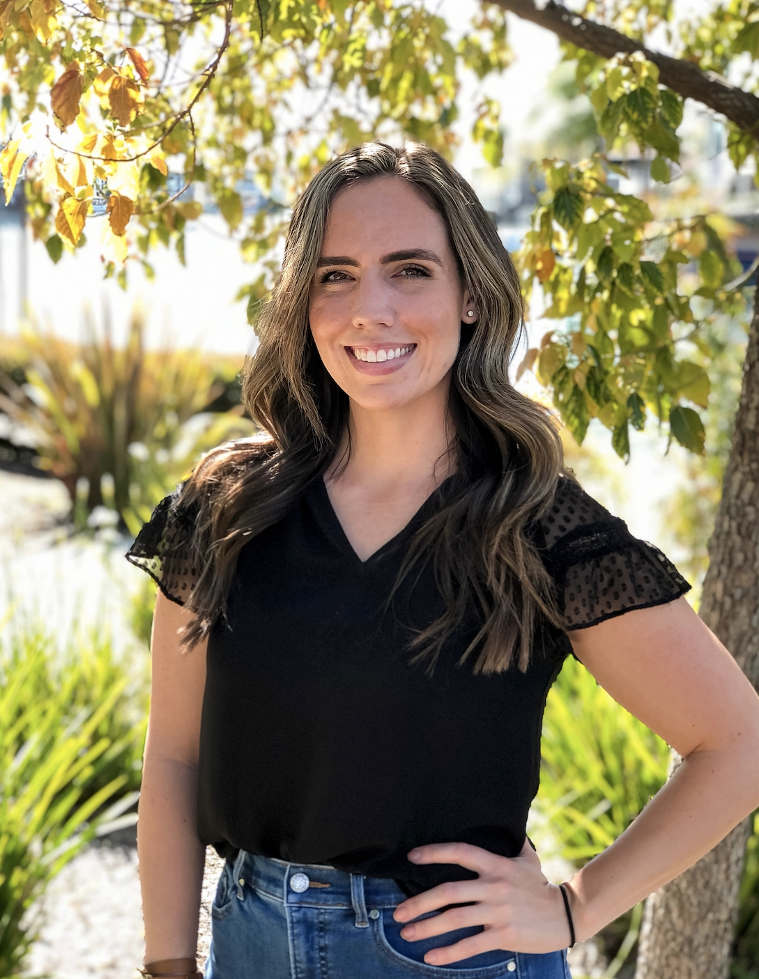 Chelsea Burch, AMFT, smiling outdoors next to a tree with green and yellow leaves, wearing a black top and blue jeans.