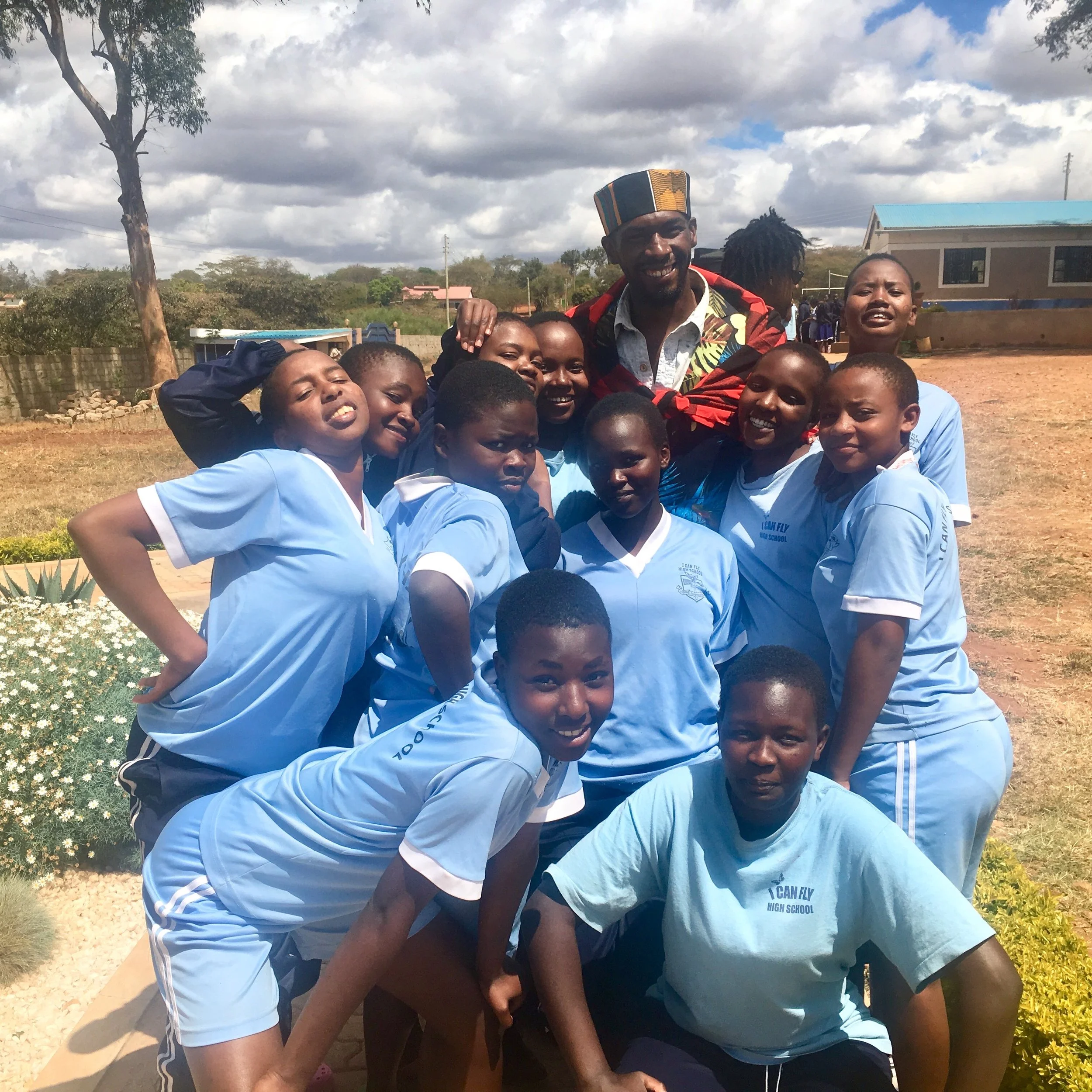 Group of children in light blue sports uniforms and a man in colorful clothing posing outdoors on a sunny day.