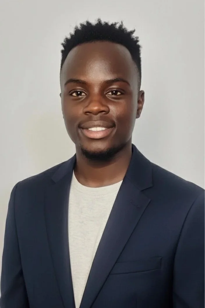 A young man dressed in a dark blazer and light shirt, smiling against a plain background.