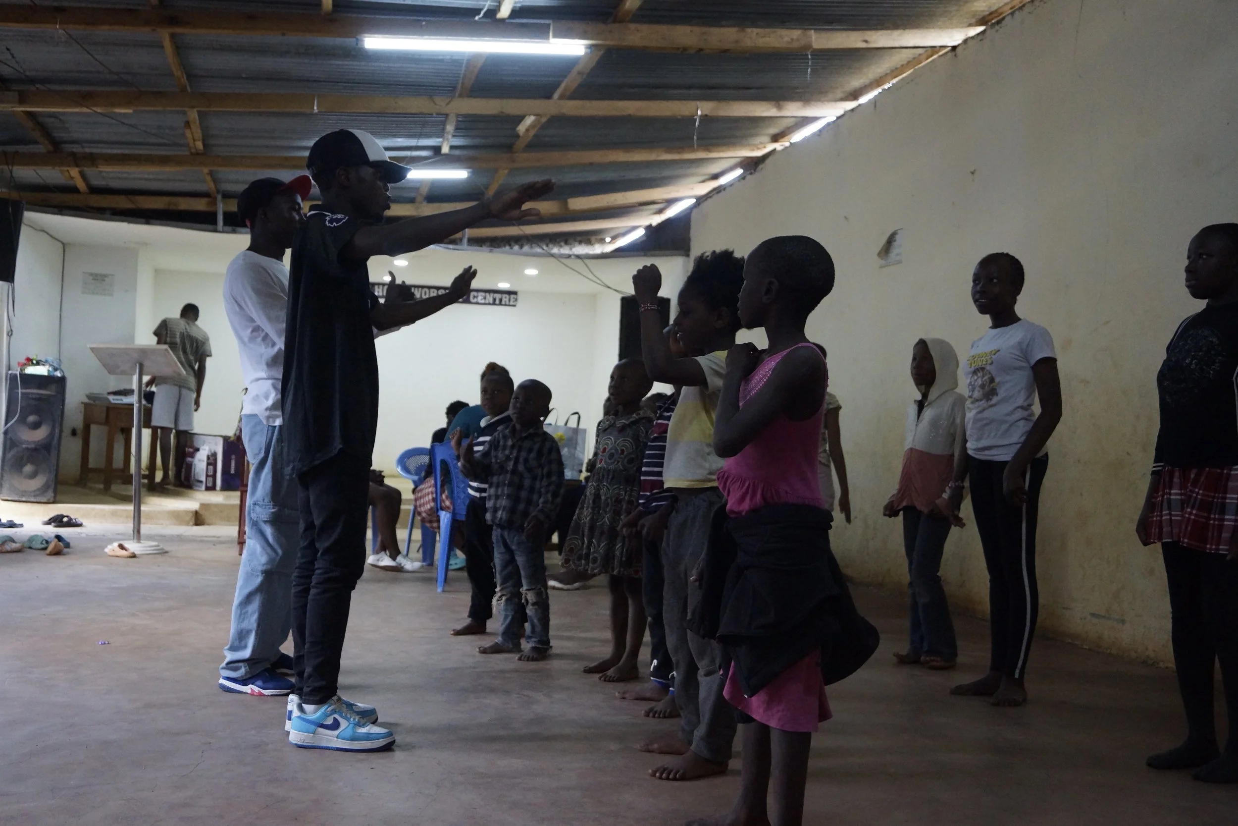 Group of children standing in a line inside a community center while a man leads them in an activity. Adults sit on chairs in the background, and another person stands near a podium.