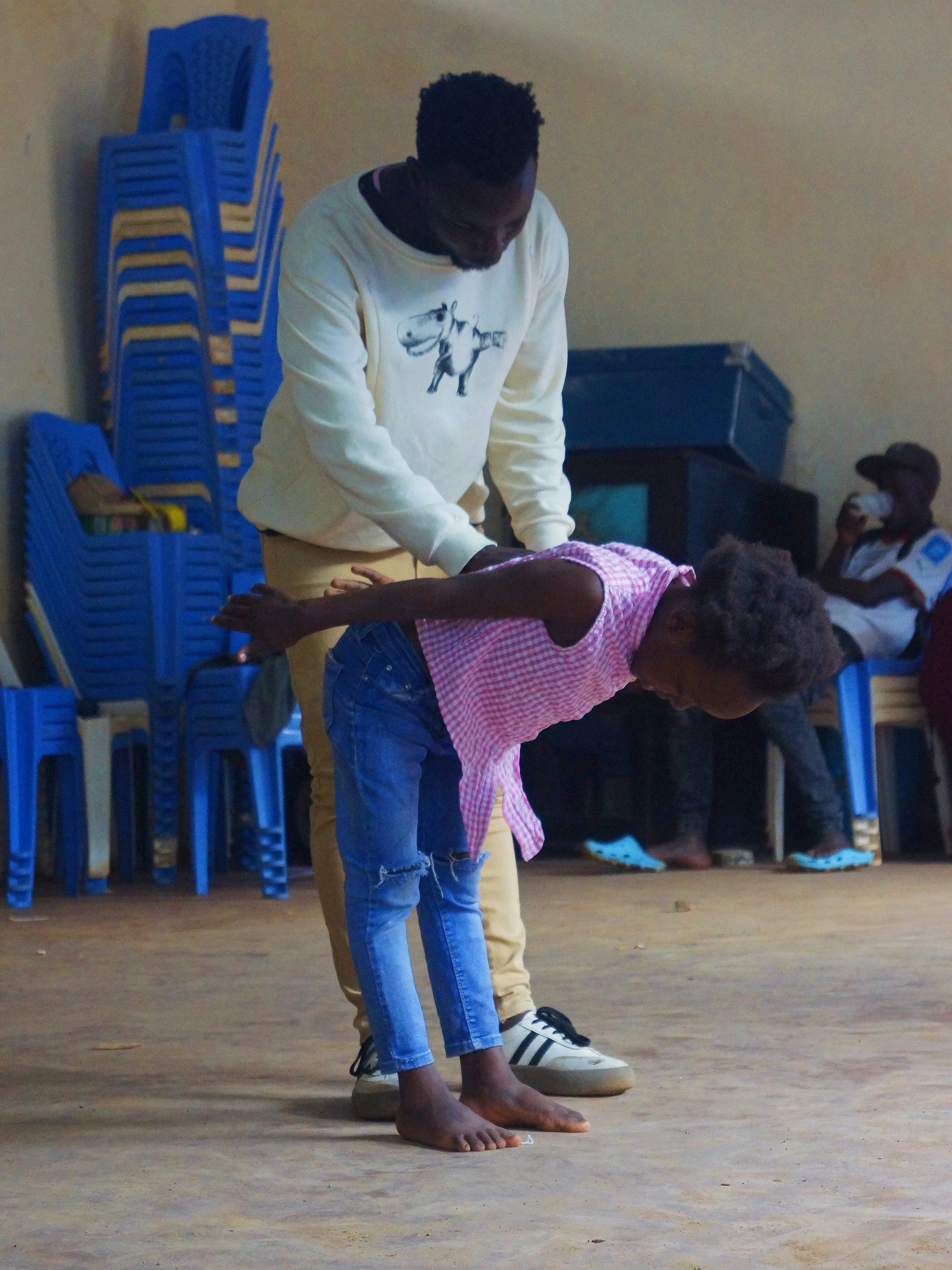 A man helping a young girl practice a dance move in an indoor setting, with chairs stacked against the wall in the background.