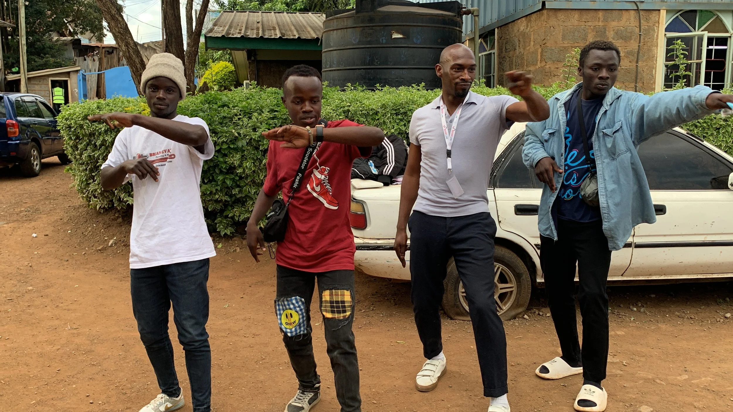 Four young men standing outdoors on a dirt ground, posing with their right arms extended in a martial arts stance. Behind them are a bush, parked cars, and a building with blue-tinted windows.