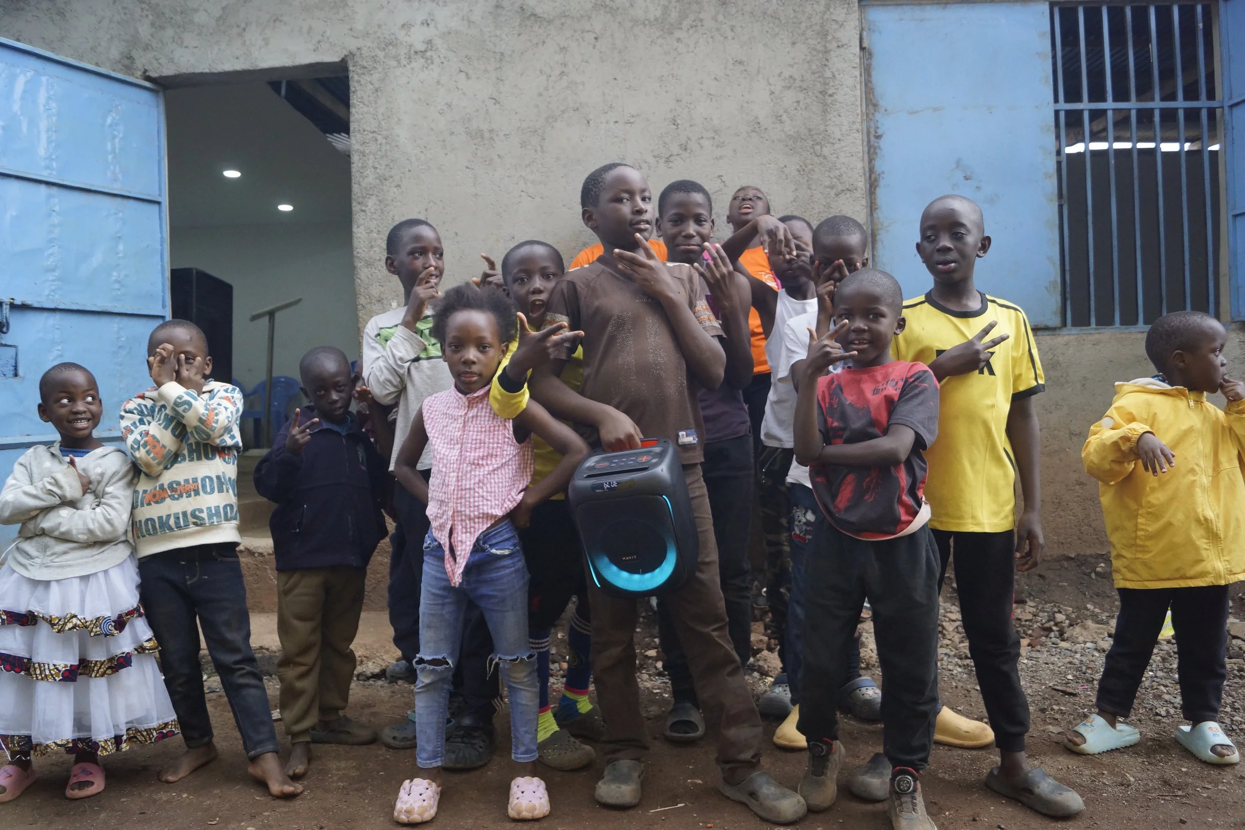 A group of children standing outside in front of a building with an open door and barred window. Some children are posing and smiling, with a few making peace signs or gestures. The ground is dirt and some children are barefoot or wearing sandals.